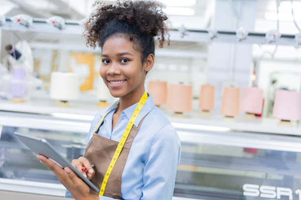 一位女设计师的特写照片是一位女裁缝，笑容灿烂，手里拿着写字板和笔，从事服装设计。织造工业用卷尺。旁边有一台缝纫机在工作。图片下载