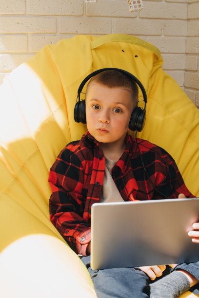 一个戴着黑色耳机的学龄男孩坐在一个黄色的豆袋上，手里拿着一台平板电脑。孩子和小玩意图片下载