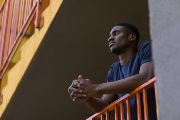 非裔美国年轻人站在阳台上，在楼梯上向外看。男子把手放在黄色大楼的橙色栏杆上。水平面。文本复制空间图片下载