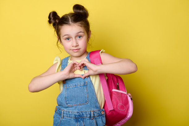 迷人的女学生带着粉红色的书包，用手指做心形，对着黄色背景上孤立的相机微笑图片下载