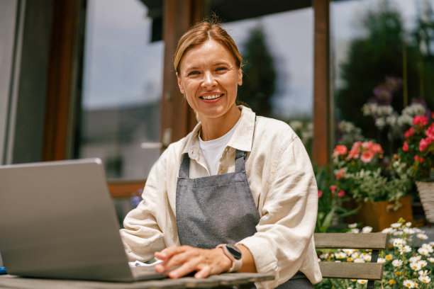 微笑的花商在笔记本电脑上工作，看着相机在商店里接受网上订单图片下载