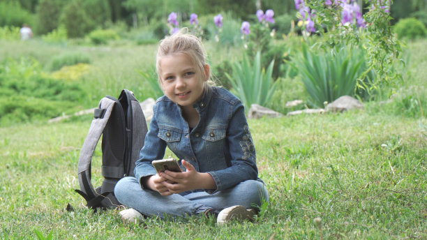 一个沉思的九岁女孩，一个女学生，坐在公园的草地上。她手里拿着智能手机，沉思着。图片下载