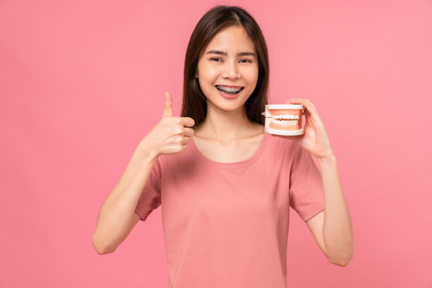 微笑的亚洲女性戴着牙套，竖起大拇指，拿着粉色背景上的牙齿模型，口腔卫生和保健概念。图片下载