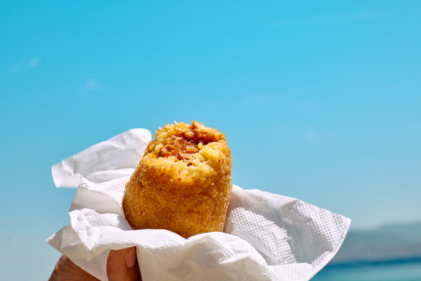 典型的西西里街头小吃在海边背景。沙滩上的男人手里拿着热腾腾的美味arancina(带肉的油炸饭团)。外卖食品。图片下载