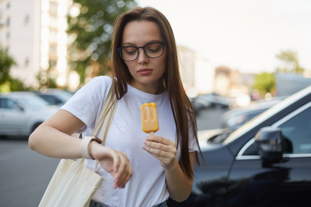 戴着眼镜的年轻美女一边吃着黄色冰淇淋一边查看智能手表上的通知。自然的女性，棕色长发，使用智能手表小工具图片下载
