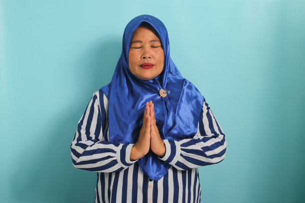 一位戴着蓝色头巾的亚洲中年妇女闭着眼睛祈祷，独自在蓝色背景下。图片下载