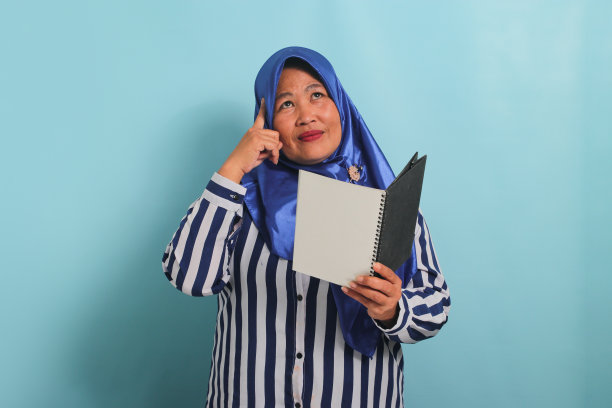 戴着蓝色头巾的体贴的亚洲妇女拿着打开的书，沉思着，蓝色的背景。图片下载