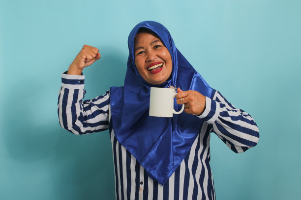 兴奋的亚洲妇女戴着蓝色头巾，举起手臂，弯曲肌肉，端着咖啡。隔离在蓝色区域。图片下载