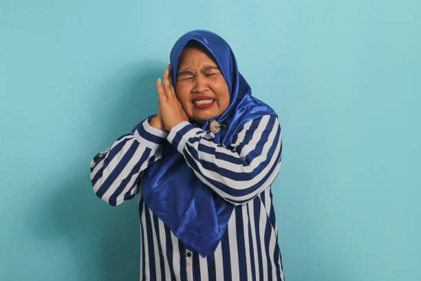 一名戴着头巾的亚洲女性因蓝色背景的噪音而感到耳朵疼痛，她捂着耳朵。图片下载