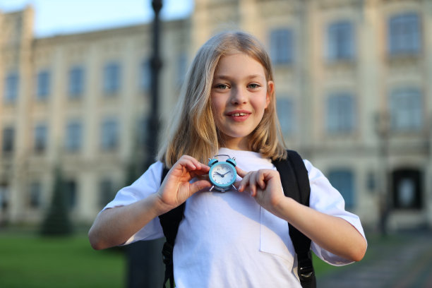 微笑的可爱的一年级女孩手里拿着一个背包和一个钟。图片下载