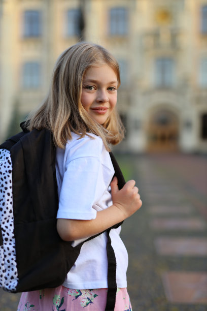 现代的一年级小女孩背着双肩包在学校的背景上。图片下载