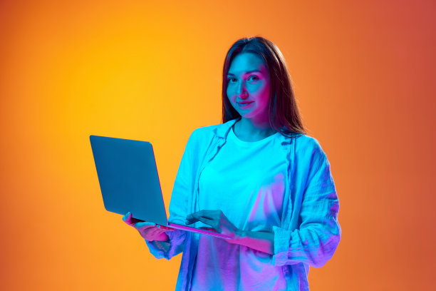 在霓虹灯的渐变橙色工作室背景下，穿着休闲服的年轻女子站在笔记本电脑上。网上购物图片下载
