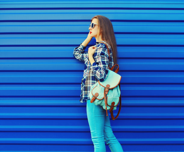 年轻女子与背包看别处穿着衬衫，太阳镜在蓝色的背景图片下载