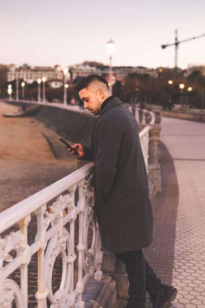 日落时分，在多诺斯塔-圣塞巴斯蒂安海滨散步，一名年轻的白人男子在使用智能手机。图片下载
