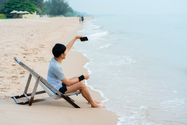 一个英俊的亚洲男人，坐在海边的椅子上自拍，海浪拍打着海岸，开心地看着他，一个受欢迎的旅游目的地。图片下载