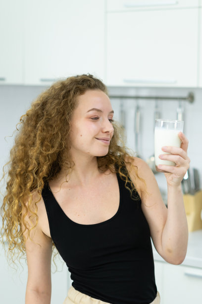 美丽的白种人女人微笑着，喝着牛奶，站着，看着厨房里的牛奶杯。美丽的白人妇女烹饪健康饮食水果或蔬菜在厨房图片下载