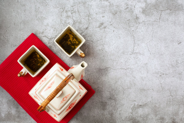 灰色背景的红色毛巾上放着一个中国风格的方形茶壶和两个茶杯。图片下载