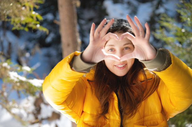 迷人的白人年轻女子显示心的手势和通过它微笑的冬季森林。一个穿着温暖冬衣的女孩祝大家都爱图片下载