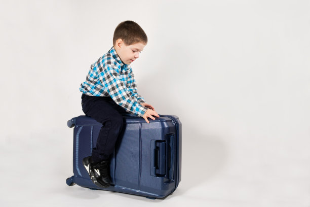 一个4岁的男孩坐在白色背景下的蓝色旅行箱上，上面留有文字空间。图片下载