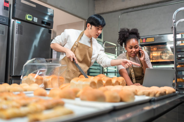 年轻的亚洲男性和非洲女性穿着围裙，在工作地点用笔记本电脑和客户打电话。情侣卖家在网上浏览手机。经营理念。零售行业。图片下载