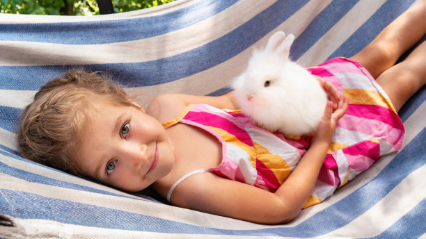 一个可爱的女孩抱着一只毛茸茸的白兔。在一个阳光明媚的日子里，孩子抱着一只装饰性的兔子坐在吊床上。快乐的小女孩带着积极的情绪对她的宠物微笑。图片下载