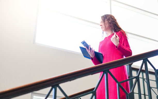 一名女商人穿着鲜艳的裙子，站在灯光明亮的办公大楼的楼梯上，手里拿着文书和手机，同时处理多项任务图片下载