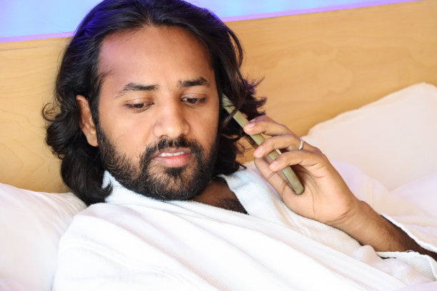 特写照片:一名印度男子身穿白色酒店毛巾长袍，斜倚在床上，靠在枕头上，把智能手机举到耳边，聚焦在前景图片下载