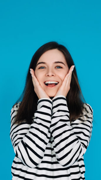 一个惊讶和快乐的年轻女子的富有表现力的肖像，手掌在颧骨上，孤立的蓝色背景图片下载