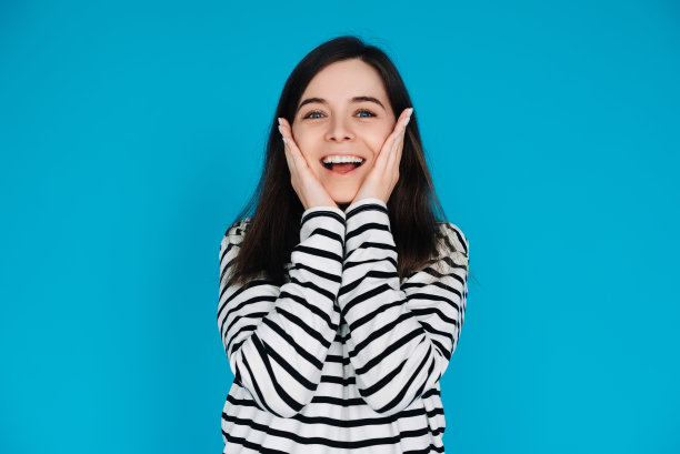 一个惊讶和快乐的年轻女子的富有表现力的肖像，手掌在颧骨上，孤立的蓝色背景图片下载