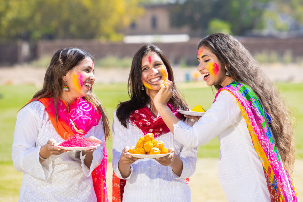 一群快乐的年轻印度妇女穿着白色的kurta dupatta服装，在公园庆祝胡里节，手里拿着装满gulal和laddoo糖果的盘子，妇女们玩着五颜六色的粉末。图片下载