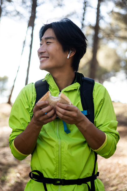 一个年轻的亚洲男子正在爬山的垂直照片。微笑的年轻人一边看一边吃三明治。自然概念，亚洲之旅。在树林里搭个帐篷。图片下载