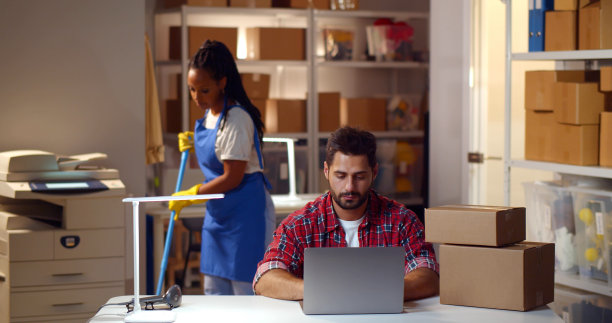 网上商店仓库经理在手提电脑前工作，背景是女看门人拖地图片下载