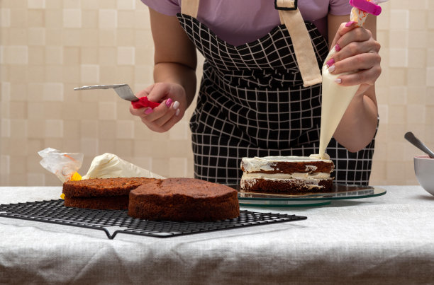 一个女人的手拿着糕点袋，把白色奶油挤到饼干上。另一只手拿着厨房铲子。前景是干净的蛋糕。有选择性的重点。图片为食品，糖果商的文章。图片下载