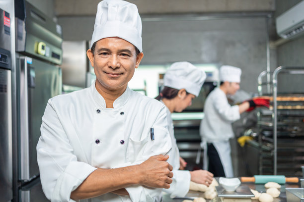 高级烘焙师看着镜头。面包师穿着厨师服，戴着厨师帽，在厨房里一起做饭。年轻的亚洲人从厨房的现代电炉中取出新鲜出炉的饼干。图片下载
