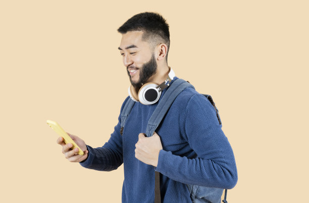 年轻微笑的亚洲男子带着耳机和手机，拿着一个背包，米色的背景图片下载