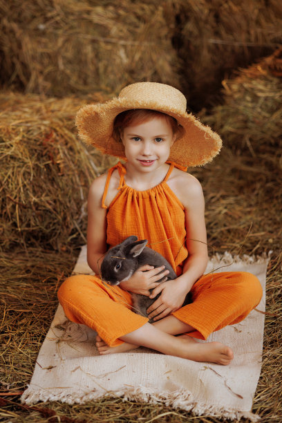 可爱的小女孩穿着橙色的裙子，戴着草帽，和一只棕色的兔子坐在稻草捆上图片下载