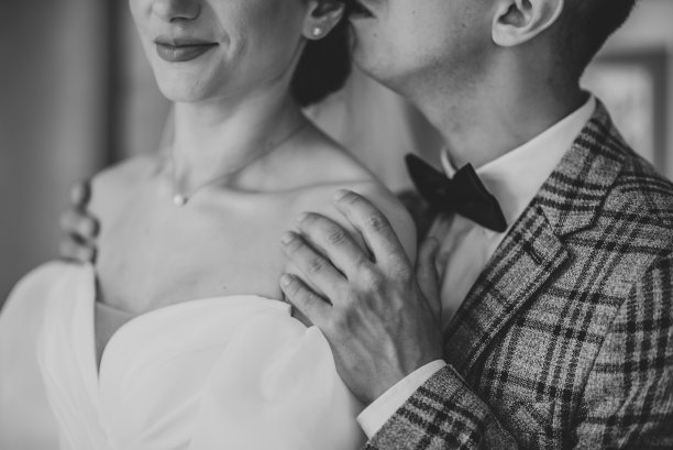 一对热恋中的情侣。新郎在新娘身后亲吻，拥抱肩膀特写。男人和女人站在一起摆姿势。在摄影棚里拍摄黑白照片。结婚的那一天。第一次见面是新婚夫妇。图片下载