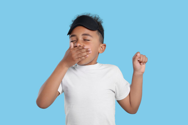 戴着睡眠面罩的男孩打着哈欠，在浅蓝色的背景下伸展身体。失眠的问题图片下载