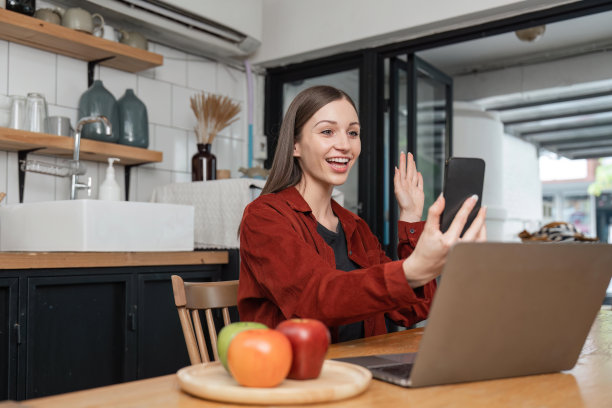 微笑着的年轻女子正在和同事进行视频通话，她坐在厨房里拿着电话，挥手图片下载