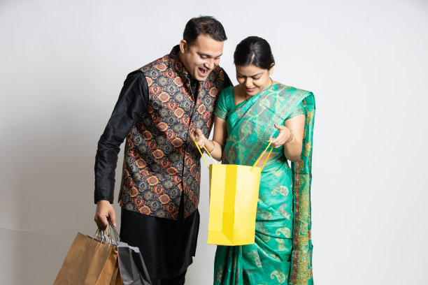 快乐惊讶的年轻印度夫妇在购物袋里找东西。惊奇的男人和女人购物者穿着民族kurta和纱丽服装站在孤立的白色背景图片下载
