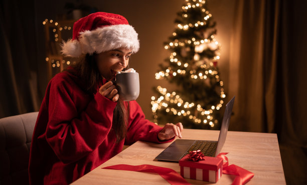 圣诞节期间，戴着圣诞帽的女人在家里喝着可可和棉花糖，看着笔记本电脑屏幕，坐在桌子前上网看电影图片下载