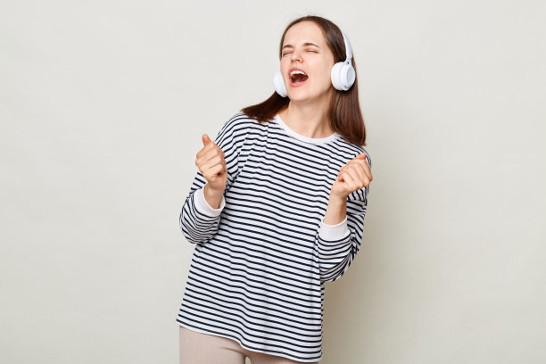 穿着条纹衬衫的年轻女子戴着耳机听着音乐，在灰色的背景下跳舞，唱着最喜欢的歌。图片下载