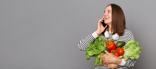 美女抱着新鲜蔬菜从超市回来，穿着条纹衬衫，隔着灰色的背景讲着手机，看着广告版面。图片下载