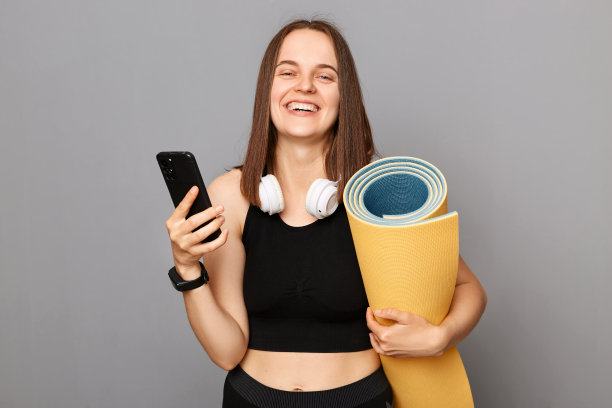 满意的女人穿着黑色运动服，拿着瑜伽垫，站在灰色的背景中，拿着手机浏览互联网，购买在线运动训练的锻炼。图片下载