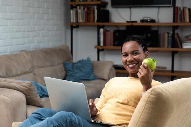 非裔美国妇女在客厅里阅读在线PDF书和吃苹果。健康生活，闲暇时间在家享受。图片下载