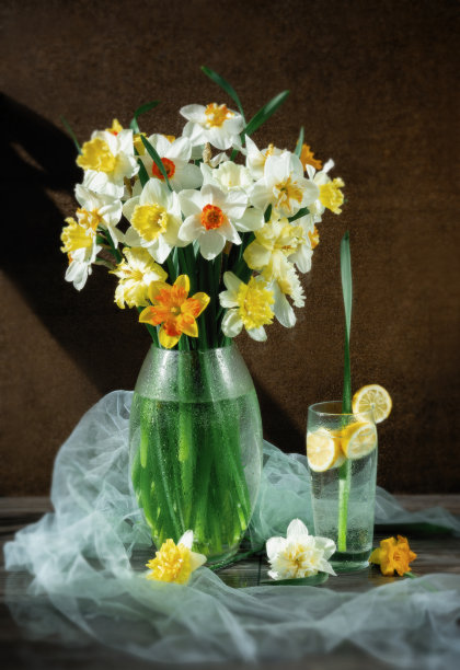 一束不同品种的黄色和白色水仙花放在玻璃花瓶里，柠檬水放在透明玻璃杯里。黑色背景上的水仙花。图片下载