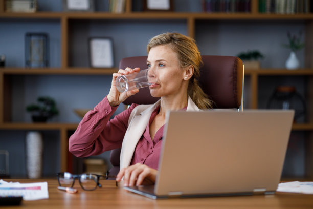 在办公室用笔记本电脑工作的疲惫的女商人。压力大的女人拿杯水喝。医疗保健和健康的生活方式。饮用水。饮食的概念。健康饮食。脱水图片下载