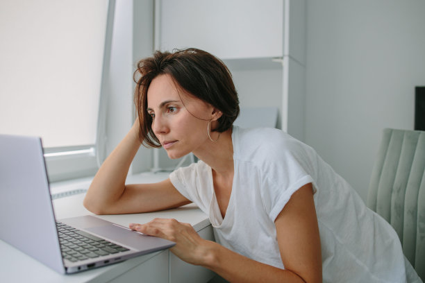 疲惫，沮丧，疲惫，压力大的女人坐在办公桌前用笔记本电脑工作。女性在白天工作。穿白t恤的女孩图片下载