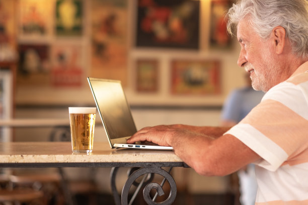 聪明的工作理念。一个面带微笑的老人在酒吧里一边用笔记本电脑工作，一边喝着新鲜的啤酒。有胡子的老男人在远处干活图片下载