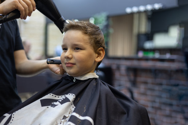 理发师用吹风机吹一个男孩头上和脖子上剪下来的头发。这个孩子在理发店剪了一个漂亮的发型。一个孩子在美容院的概念。图片下载